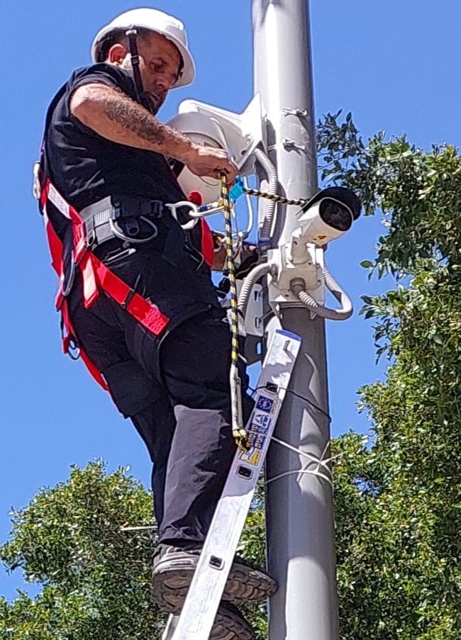 טכנאי עם רתמת בטיחות מחבר מצלמת אבטחה לעמוד מול שמיים כחולים